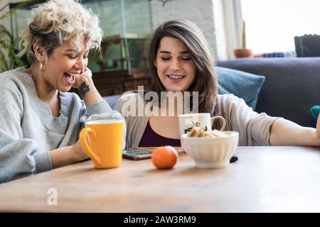 Glückliche Mitbewohner, die mit dem Smartphone unterwegs sind, während sie Kaffee am Tisch haben Stockfoto
