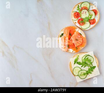 Drei köstliche Sandwiches mit Lachs, Tomaten, Gurke und Käse auf hellem Marmortisch, Kopierraum Stockfoto
