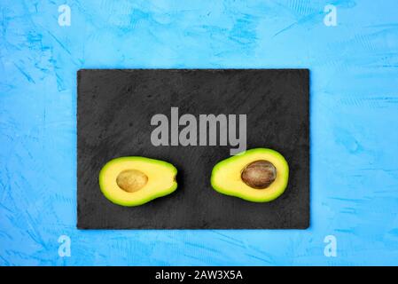 Avocado, halbiert, eine Schicht mit Kern auf grauem Schiefer sind auf einer helltürkisfarbenen Oberfläche, einer flachen Position, einem Bild mit Kopierraum angeordnet. Stockfoto