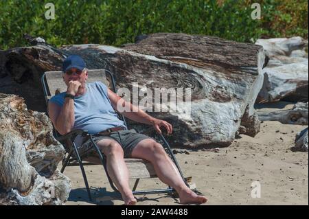 Mit einer Kugelkappe und einer Sonnenbrille eine Seniorenlounge auf einer Klappchaise in der Nähe von großem Treibholz am Strand im Siletz Bay Park in Lincoln City, Oregon Stockfoto