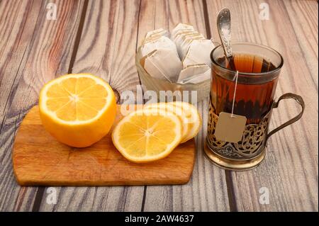 Tee aus facettiertem Glas in einem Vintage-Cup-Halter, Teebeutel in einer Glasvase und geschnittene Zitrone auf einem Brett auf Holzhintergrund. Nahaufnahme Stockfoto