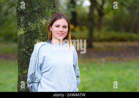 Schönes glückliches Mädchen im herbstlichen Park, Nahaufnahme des Porträts der süßen blonden Frau mit stilvollem Beret, attraktive Frau umarmende Baumstamm in schöner Lage Stockfoto