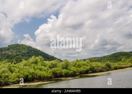 Blick vom ruhigen Fluss auf grüne Bäume und Büsche vor dem Hintergrund hoher felsiger Berge inmitten eines sonnigen Tages. Große weiße Wolken in Stockfoto