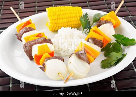 Gegrilltes Sirloin-Steak und Gemüse-Schisch-Kebabs, serviert mit Mais auf dem Cob und weißem Reis. Hintergrund ist eine Bambusplatzmatte. Stockfoto