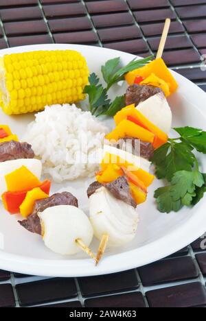 Gegrilltes Sirloin-Steak und Gemüse-Schisch-Kebabs, serviert mit Mais auf dem Cob und weißem Reis. Hintergrund ist eine Bambusplatzmatte. Stockfoto