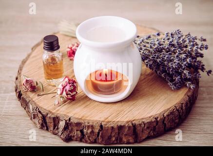 Vintage-Style-Bild der weißen Kerze Aroma Öllampe mit ätherischer Ölflasche und trockenen Blumenblättern auf naturbelassener Kiefernholzscheibe, trockener Hintergrund Stockfoto