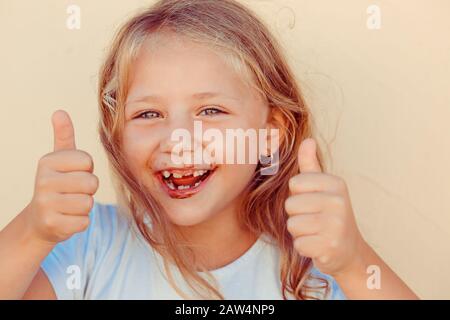 Lecker, ich mag es. Das kleine Mädchen, das gerade einen Kuchen aus Schokolade aß, der mit den Händen die Daumen nach oben zeigt, lockig blonde gebräunte Kind. Lustige pi Stockfoto