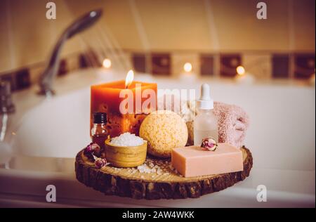 Home Spa-Produkte auf Holztablett: Seifenbarsch, Badbombe, Aromabadsalz, ätherische und Massageöle, Kerze brennen, rollendes Handtuch im Badezimmer vorbei Stockfoto