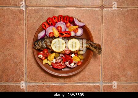 Gegrillte Forelle mit Gemüse, roten und gelben Kirschtomaten, Pfeffer, roter Zwiebel, Oliven, Zitrone und Gewürzen in rustikaler Keramikplatte auf altem Keramiktabl Stockfoto