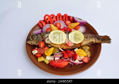 Gegrillte Forelle mit Gemüse, roten und gelben Kirschtomaten, Pfeffer, roter Zwiebel, Oliven, Zitrone und Gewürzen auf rustikaler Keramikplatte über weißem Backgrou Stockfoto