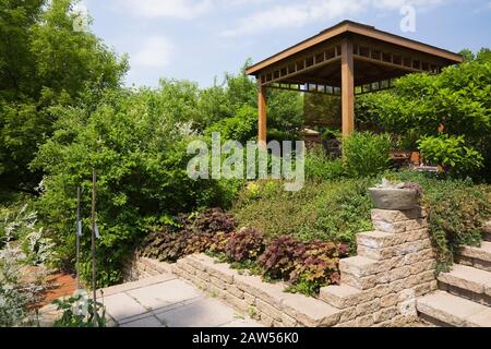 Erhöhte Steingrenze zu Heuchera - Korallenblumen und braune Holzgazebo im privaten Hinterhofgarten im späten Frühjahr. Stockfoto