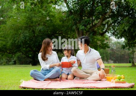 Asiatische Teenager Familie fröhlicher Urlaub Picknick-Moment im Park mit Vater, Mutter und Tochter Lesen Buch und Lächeln zu verbringen Urlaub glücklich zusammen Stockfoto