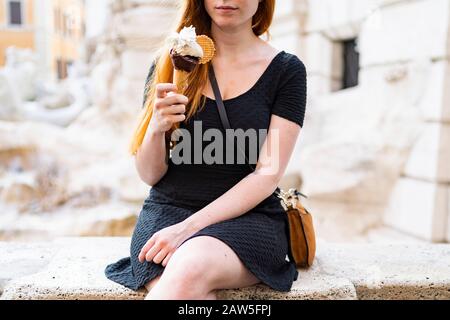 Ernten Sie junge legere Frauen mit roten Haaren, die Eiskegel halten, während sie auf dem Steinzaun sitzen und im heißen Sommertag auf der Straße der Stadt ruhen Stockfoto