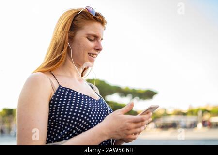 Fröhliche Ingwer-Frau in Ohrhörern lächelnd und im Smartphone beim Musikhören am sonnigen Sommertag Stockfoto