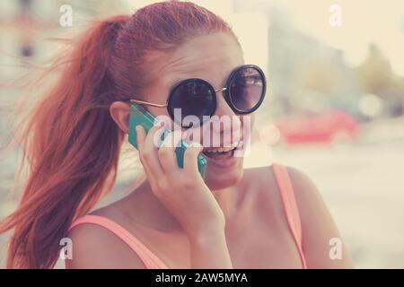 Portrait überraschte lachende, schreiende Frau, die im Sommer telefonisch auf dem Hintergrund der Stadt spricht Stockfoto