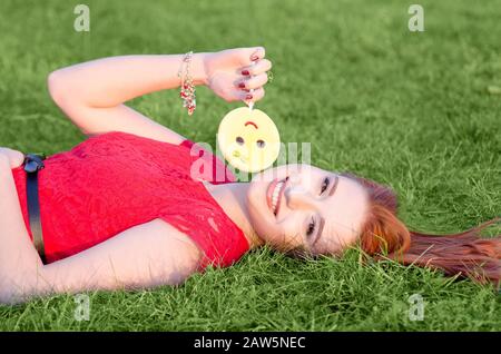 Süßes junges Rotkopfmädchen mit gelb gefärbtem Lollipop lächelt im Sommer im warmen Park auf grünem Gras liegend Stockfoto