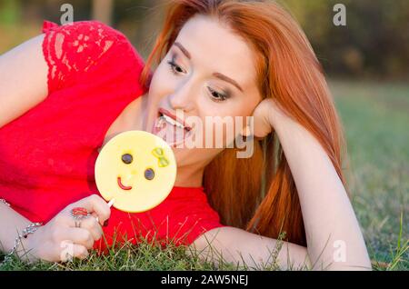 Lust auf Zucker und Emotionen. Schönes und redseliges glückliches Mädchen in einem roten Kleid, das eine lollipop Emoticon-Süßigkeit beißt, die auf Gras liegt Stockfoto