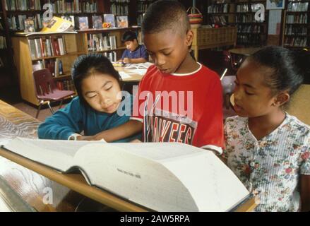Multiethnische Gruppen von Grundschulschülern sehen sich das ungekürzte Wörterbuch in der Schulbibliothek an. Stockfoto