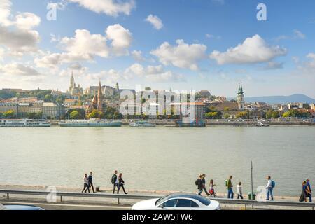 Fußgänger wandern am Ufer der Donau in Budapest, Ungarn, mit dem Buda-Schlosskomplex und der St.-Annen-Kirche über den Fluss. Stockfoto