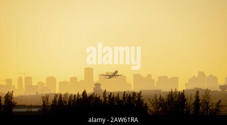 Beim Start vom Flughafen Miami mit der Stadt im Hintergrund wird ein Flugzeug vor einem farbenfrohen Sonnenaufgang in Gelb und Orange silhouettiert. Stockfoto