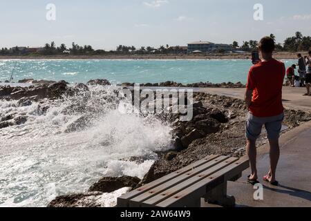 Ein Mann fotografiert das Wasser mit seinem Handy, während die Brandung die Felsen im Jetty Park am Fort Pierce Inlet an Floridas Treasure Coast abstürzt. Stockfoto