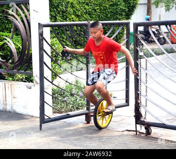 Ein junger asiatischer Junge im Alter von 12 bis 13 Jahren übt das Reiten eines Monokycle aus und hält zunächst an einem Metalltor fest, um sich selbst zu stützen, in Bangkok, Thailand, Südostasien. Stockfoto