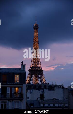 Der Eiffelturm wird bei Sonnenuntergang von Lichtern beleuchtet, mit rosa himmelblauen Wolken und goldenen Lichtern, Dachfenstern und Pariser Dächern im Vordergrund Stockfoto
