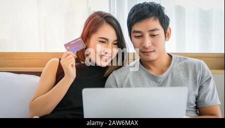 Asiatische Paare kaufen online und bezahlen mit Kreditkarte am Laptop, Glückliches Paar zu Hause Surfen im Internet im Bett Stockfoto