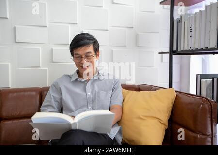 Senior Asian man liest Buch auf dem Sofa im Wohnzimmer zu Hause, Portrait of Asian Altern man ist Entspannend und Glücklich Mit Read a Magazine Stockfoto