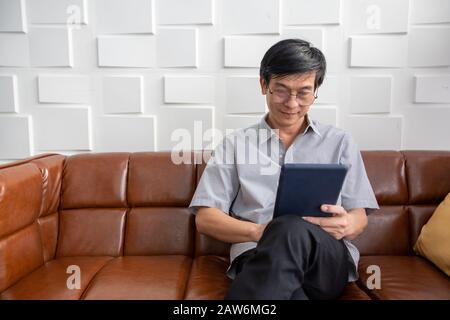 Senior Asian man spielt Tablet auf dem Sofa im Wohnzimmer zu Hause Portrait des asiatischen älteren Mannes ist Entspannend und Glücklich Mit spielendem Tablet Stockfoto