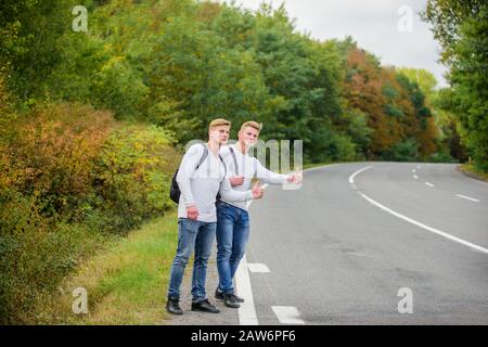 Beginnen Sie mit dem Wandern ein tolles Abenteuer in Ihrem Leben. Firmenfreunde Reisende, die auf dem Naturhintergrund der Straße hitchwandern. Fahren Sie in der Nähe des Stadtrandes. Reisende auf dem Weg. Freunde, die hitchhitchhamps unterwegs Stockfoto