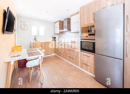 Neue, elegante, helle Küche mit Holzschränken. Geräumiges, modernes Interieur mit weißem Tisch, Stühlen, Fernseher und großen Fenstern Stockfoto