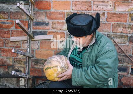 Außenporträt eines bärtigen ukrainischen Bauern, der Kürbis in die Hände nimmt und ihn betrachtet, während er gegen die Mauer sitzt Stockfoto