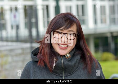 Fröhliche junge Asiatin, die in einer warmen Winteroberseite im Freien Brille trägt und dabei in urbaner Umgebung mit der Kamera lächelt Stockfoto