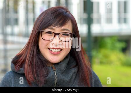 Fröhliche junge Asiatin, die in einer warmen Winteroberseite im Freien Brille trägt und dabei in urbaner Umgebung mit der Kamera lächelt Stockfoto