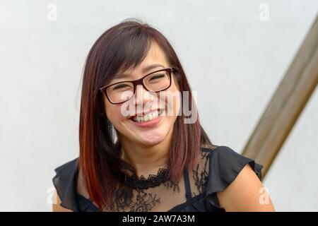 Stilvoll attraktive junge Asianerin mit Brille, die mit einem fröhlichen, freundlichen Lächeln im Freien an einer weißen Wand auf die Kamera blickt Stockfoto