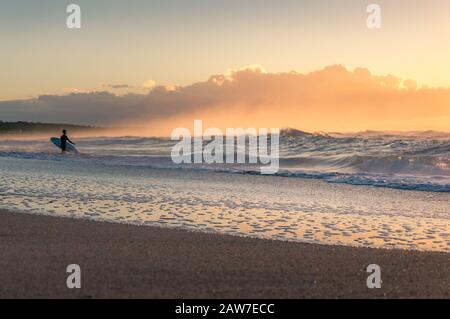 Strand Sonnenaufgang mit unerkennbarer Surferfigur in der Ferne. Sommerlicher Hintergrund am Meer. Selektive Fokussierung auf die Wasserkante Stockfoto
