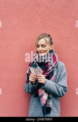 Erwachsene blond in einem Mantel und einem großen Schal gegen eine rosafarbene Wand liest eine kleine Valentinskarte Stockfoto