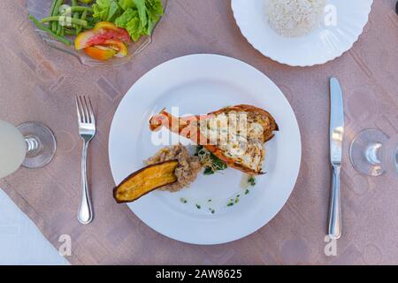 Draufsicht: Gegrillter Hummer mit frischer Tomate und Gurke auf weißem Teller, Reis und Cocktailglas auf dem Tischtuch. Stockfoto