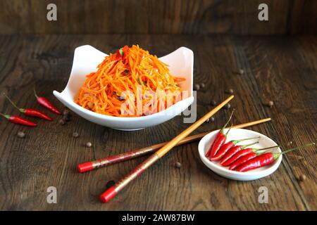 Karotten koreanischer Gemüsesalat in einer Schüssel mit Essstäbchen auf Holzeichentisch und grünen Salatblättern, frischen roten Chilischoten. Pikante Karotte im koreanischen Stil Stockfoto