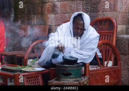 Lalibela, Äthiopien - November 2018: Mann in traditioneller weißer äthiopischer Kleidung, der Kaffeekörner bratet, bevor er einen Kaffee zubereitet. Stockfoto