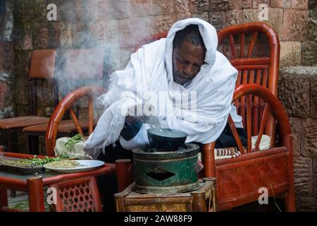 Lalibela, Äthiopien - November 2018: Mann in traditioneller weißer äthiopischer Kleidung, der Kaffeekörner bratet, bevor er einen Kaffee zubereitet. Stockfoto