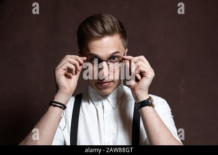 Junger Mann in runder Brille, Hosenträgern und einem weißen Hemd auf braunem Grund Stockfoto