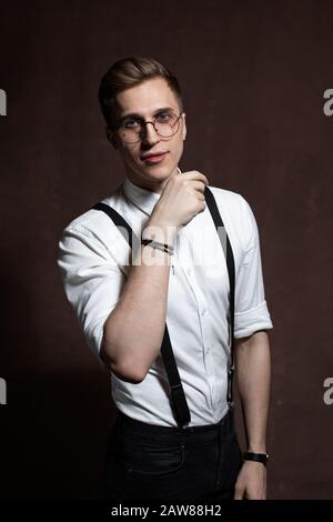 Junger Mann in runder Brille, Hosenträgern und einem weißen Hemd auf braunem Grund Stockfoto