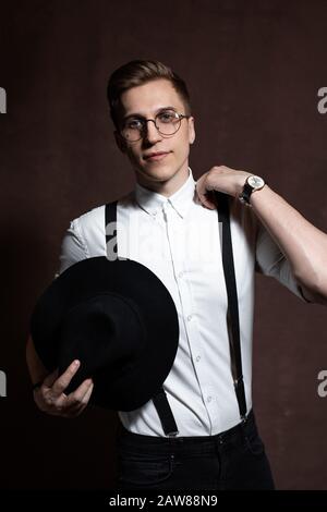Junger Mann in runder Brille, Hosenträger, weißem Hemd und Hut auf braunem Grund Stockfoto