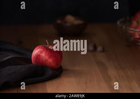 Ein heller roter apfel auf dunkelbraunem Stoff mit Nüssen im Hintergrund und einer stimmungsvollen Beleuchtung Stockfoto