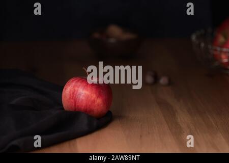 Ein heller roter apfel auf dunkelbraunem Stoff mit Nüssen im Hintergrund und einer stimmungsvollen Beleuchtung Stockfoto