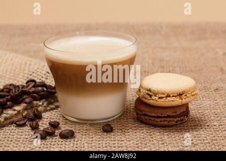 Tasse Cappuccino, Kaffeebohnen mit Leinwandsack und Makronen auf dem Hintergrund des Sacktuchs. Stockfoto