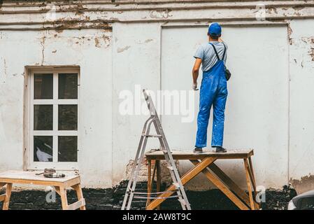 Nicht erkennbarer Arbeiter in blauer Bauhose steht auf einem Holzständer und arbeitet mit einem Spachtel mit Gips an der Wand. Renovierung der Stockfoto