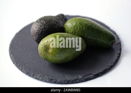 Drei grüne Avocados auf einem schwarzen Brett auf weißem Hintergrund. Rohe ganze Avocados auf dem Brett. Grüner Avocado und mit einer dunkleren Haut. Stockfoto